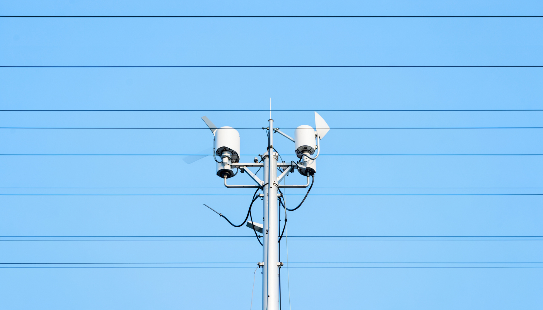 A clean architectural photograph of a weather monitoring station with strong symmetry, its white instruments and anemometer silhouetted against a pale blue sky, framed by neat horizontal lines.