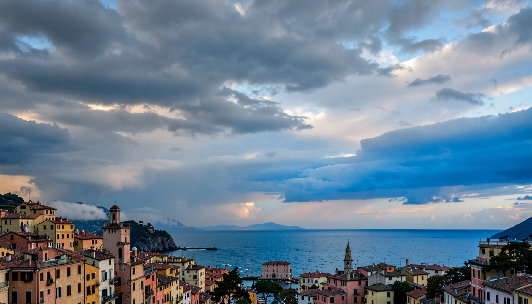 A dramatic sky over a coastal Italian town, with layered clouds in shades of slate gray and pale silver catching the last light of dusk, conveying the tension of changing weather.