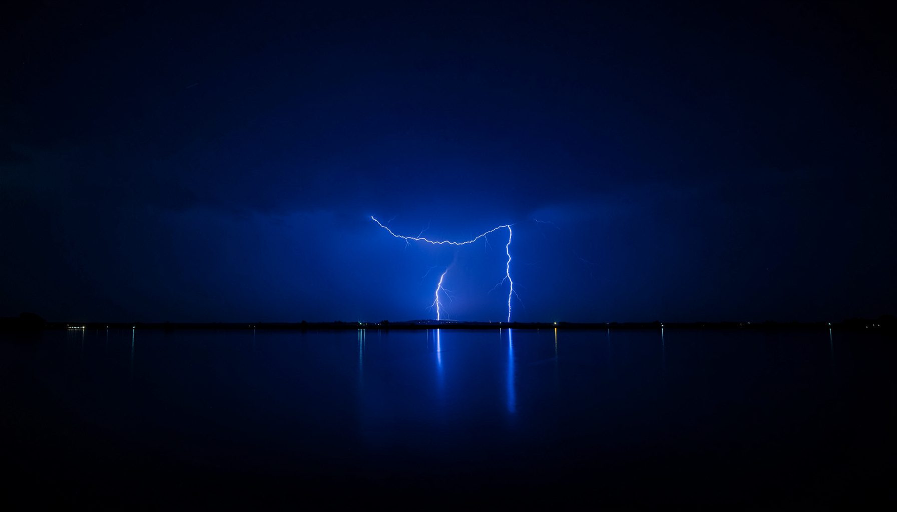 Wide dark sky with distant lightning reflected symmetrically over still water, navy blue tones, dramatic and atmospheric